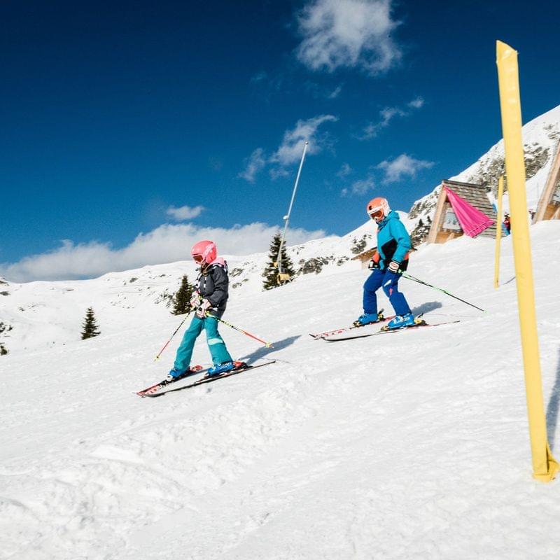 Two children wearing helmets skiing downhill on a sunny Alpine slope